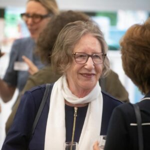 Smiling woman with short gray hair and glasses wearing a white scarf, talking at a social gathering with people in the background, holding a drink.