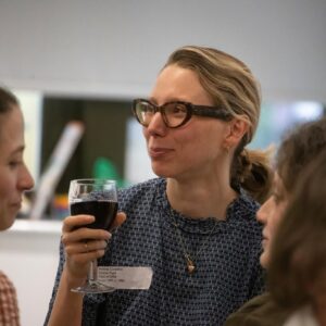 Woman wearing glasses holding a glass of red wine, chatting at a social gathering.