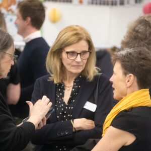 Center-right woman with blonde hair and glasses in a navy blazer talks with others at a social/business event, arms crossed.