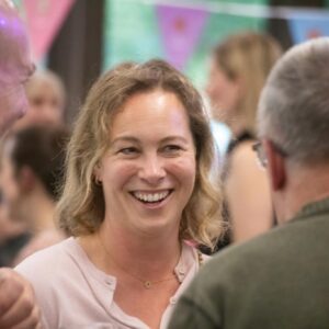 Smiling woman with blonde hair in a pink top talking with people at a crowded social gathering, colorful decorations in the background.