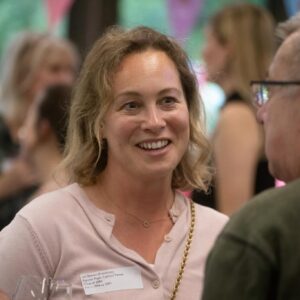 Smiling woman with blonde hair in a light pink top at a busy social event, wearing a gold chain necklace and a visible name tag on her chest.