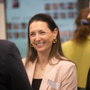 Smiling woman in a pale pink blazer with gold hoop earrings, talking with others at a group meeting.