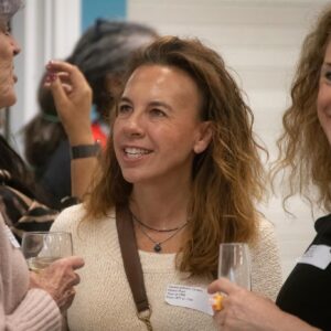 Three women in a social setting smile and chat, one holding a glass of white wine in the foreground.
