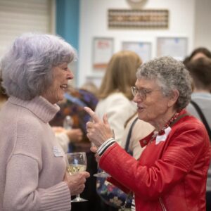 Two elderly women share a conversation at a social event, one holding a glass of white wine and wearing a light sweater, the other in a red jacket with a name badge.