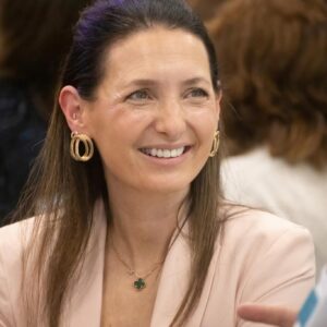 Smiling woman wearing a pink blouse with gold hoop earrings and a clover pendant necklace in an indoor setting.