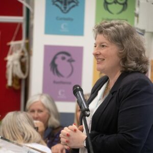 Woman in a blazer speaking into a microphone at a podium during a community event, with colorful posters on the wall behind her.