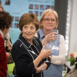 Smiling woman in a dark outfit holding a wine glass, surrounded by other guests at a social reception.