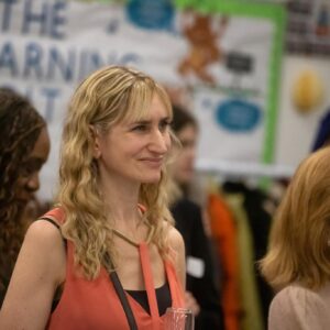 Woman with wavy blonde hair in a coral sleeveless dress at a social event, a wine glass visible in the foreground
