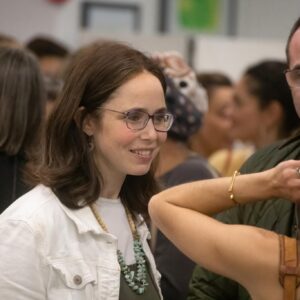 Smiling woman with glasses in a white jacket at a social gathering, being helped with jewelry by someone off to the right.