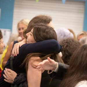 Two people share a warm hug at a crowded indoor gathering, smiling faces in the background.