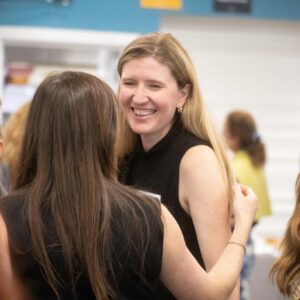 Smiling blonde woman in a black sleeveless top hugs someone at an indoor social event.