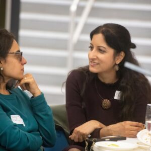 Two women chat at a table during a meal, with plates and drinks in front of them.