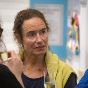 Midshot of a woman with brown hair in a yellow jacket talking to a person in blue at a social gathering, holding a champagne flute in the foreground.