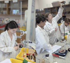 Three students in lab coats and goggles conduct chemistry experiments; one pours from a flask while others observe and take notes.