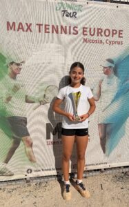 Young girl in white shirt and black shorts holding a trophy, posing in front of a tennis banner at Max Tennis Europe event in Nicosia, Cyprus.