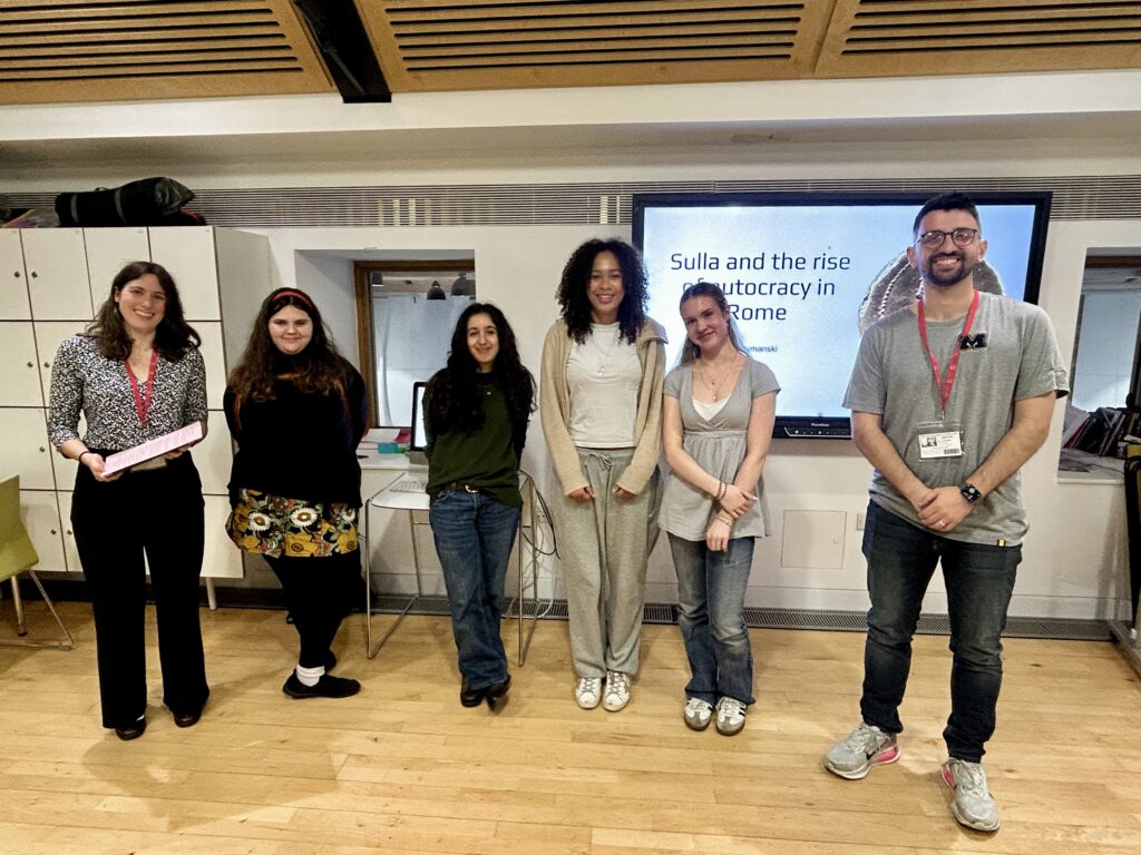 Group of six students standing in a classroom in front of a presentation screen displaying 'Sulla and the rise of autocracy in Rome'.