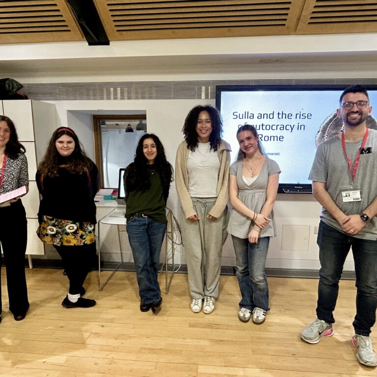 Group of six students standing in a classroom in front of a presentation screen displaying 'Sulla and the rise of autocracy in Rome'.