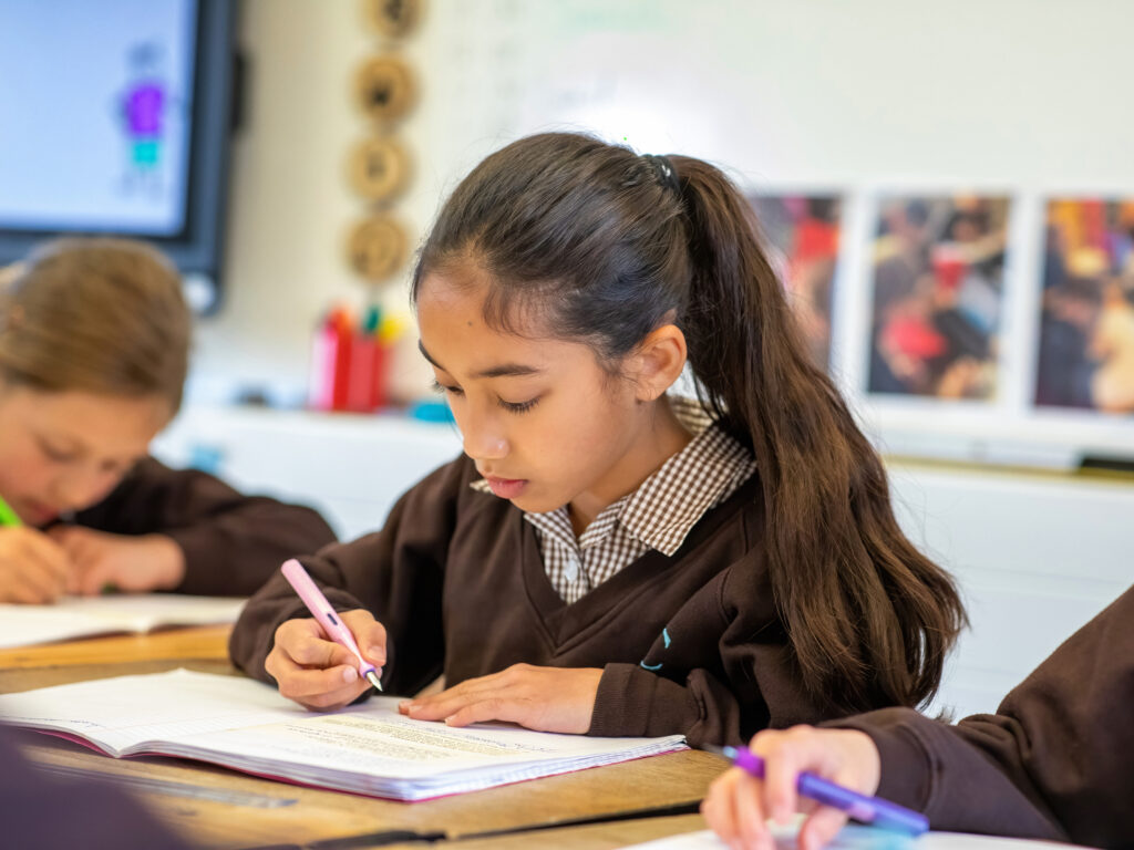 Girl with long dark hair tied back, focused on writing in a notebook at a desk in a classroom.