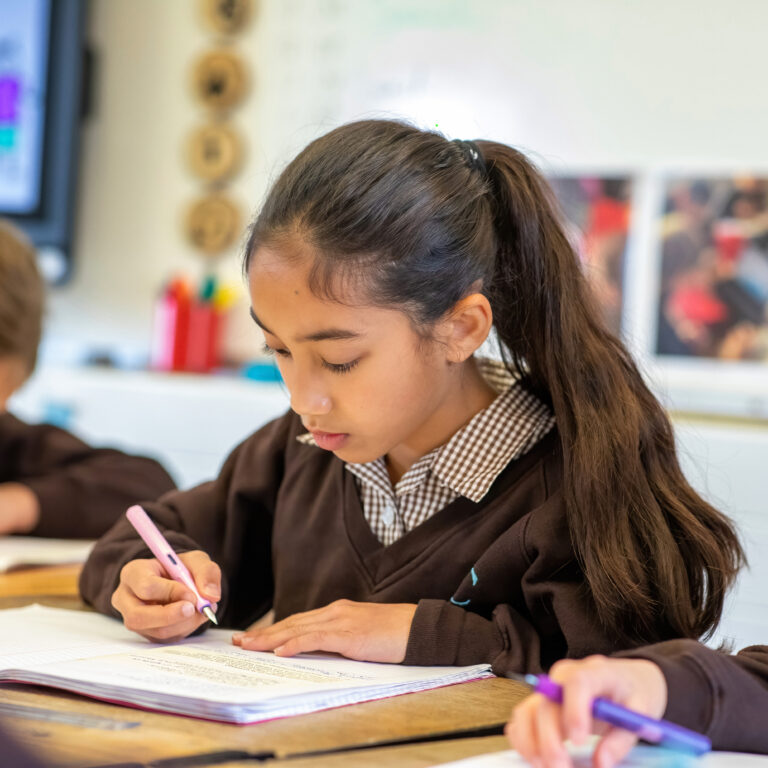 Girl with long dark hair tied back, focused on writing in a notebook at a desk in a classroom.