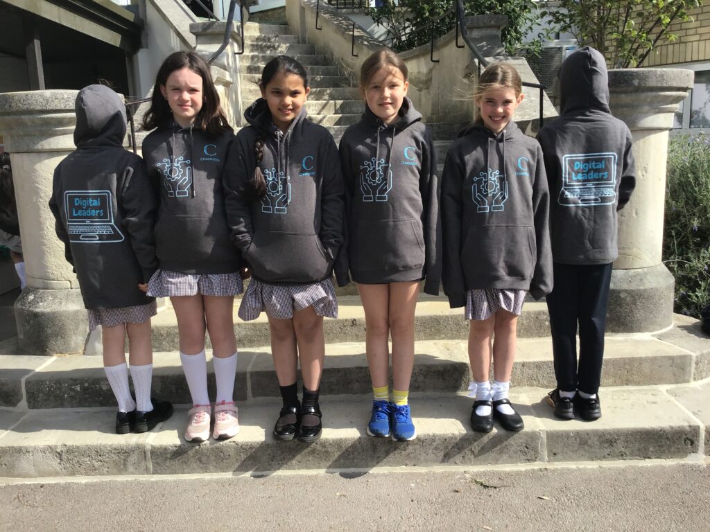 Five students stand on stone steps outdoors, wearing matching dark hoodies with a 'Digital Leaders' logo on the back and a gear/hand design on the front; some face the camera, others have their backs turned.