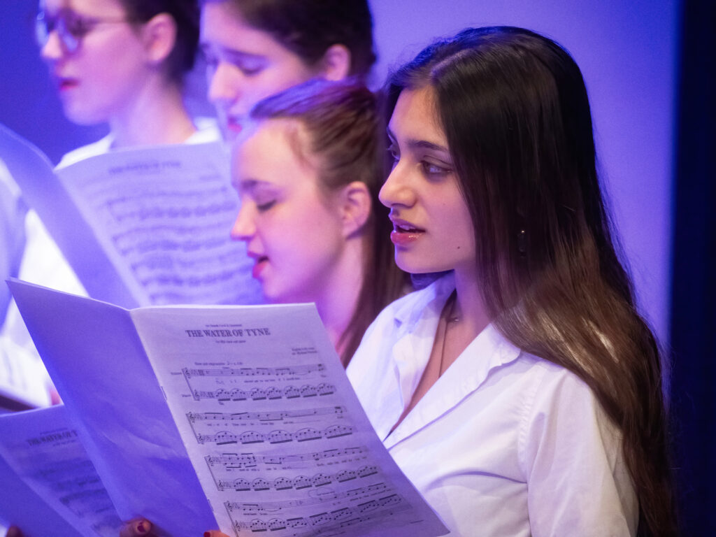 A young woman singing from sheet music under blue stage lighting, part of a choir group in white shirts.