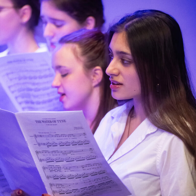 A young woman singing from sheet music under blue stage lighting, part of a choir group in white shirts.