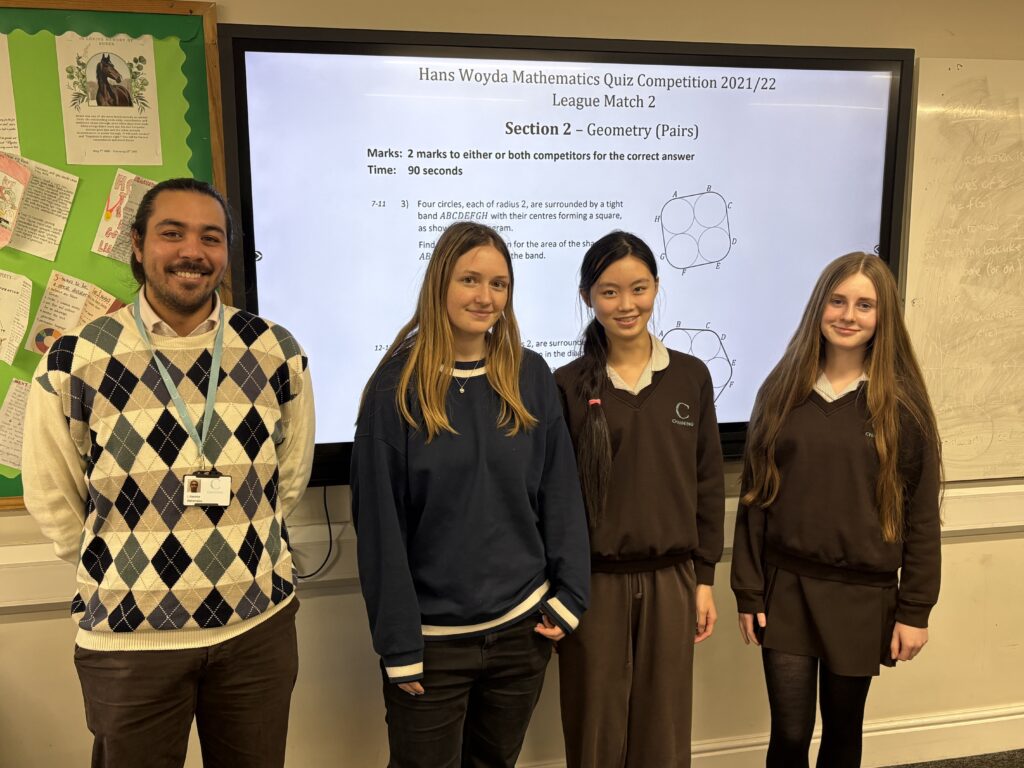 Four students stand in a classroom in front of a large screen showing a math quiz slide.