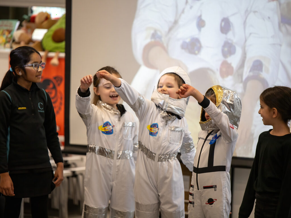 Children dressed as astronauts raise their arms in a classroom performance, wearing silver spacesuits with patches while a teacher watches from the left.