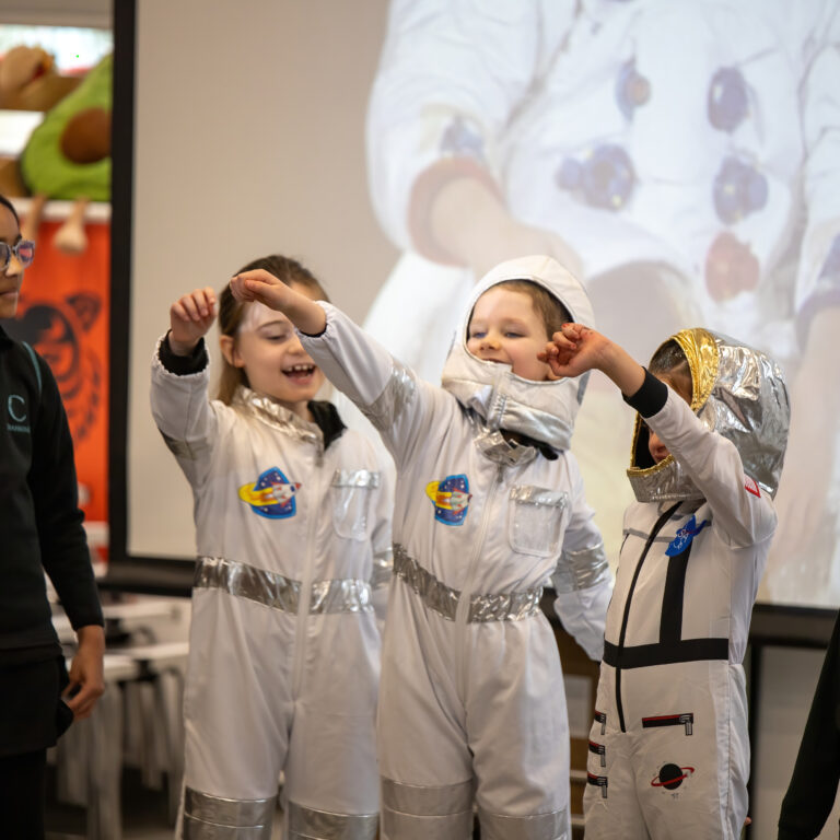 Children dressed as astronauts raise their arms in a classroom performance, wearing silver spacesuits with patches while a teacher watches from the left.