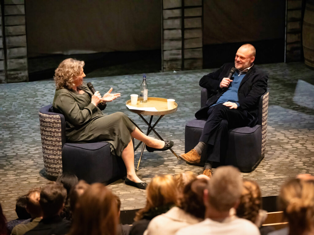 Two people sit in chairs on a stage, each holding a microphone, chatting in front of an audience with a small table between them.