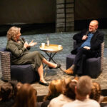 Two people sit in chairs on a stage, each holding a microphone, chatting in front of an audience with a small table between them.