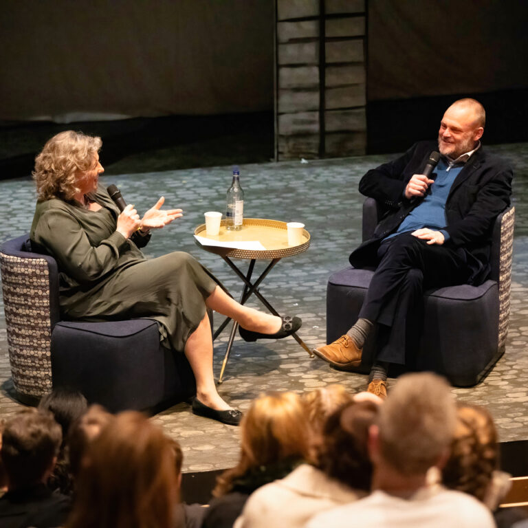 Two people sit in chairs on a stage, each holding a microphone, chatting in front of an audience with a small table between them.