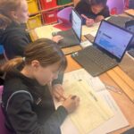 Three students seated around a table with laptops, writing in notebooks; colorful storage bins line the wall in the background.