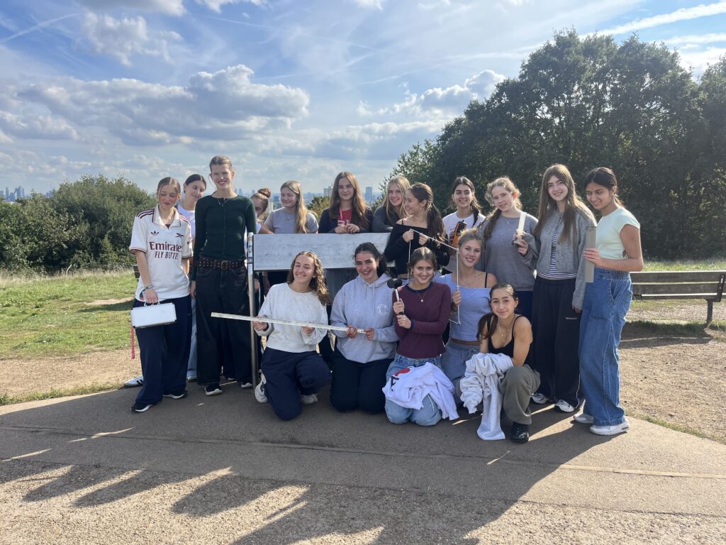 Group of teenagers posing outdoors on a sunny day, holding a long white poster and measuring tools with smiles.