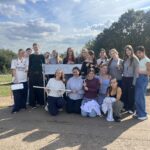 Group of teenagers posing outdoors on a sunny day, holding a long white poster and measuring tools with smiles.