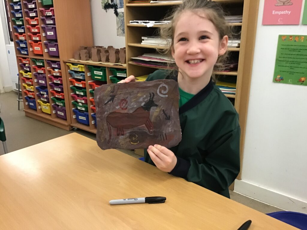 Smiling girl in a classroom holds up a brown paper artwork with animal sketches.