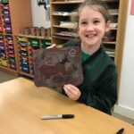Smiling girl in a classroom holds up a brown paper artwork with animal sketches.