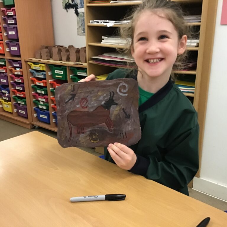 Smiling girl in a classroom holds up a brown paper artwork with animal sketches.
