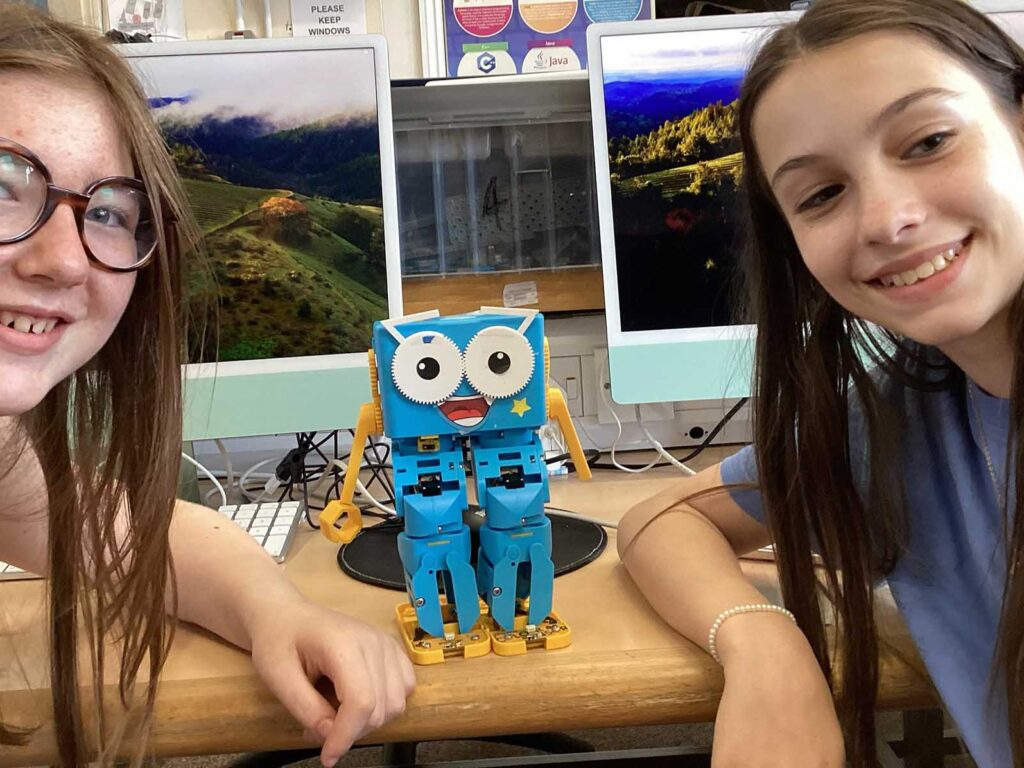 Two girls smile at the camera with a blue robot toy on the desk between them in a computer lab setting.