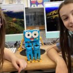 Two girls smile at the camera with a blue robot toy on the desk between them in a computer lab setting.