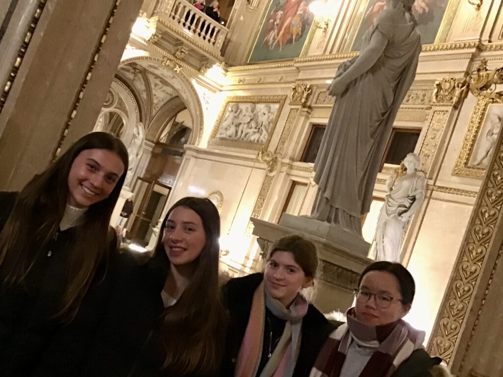Group of smiling young adults posing for a photo in a gilded, ornate museum hall with statues and murals.