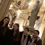 Group of smiling young adults posing for a photo in a gilded, ornate museum hall with statues and murals.