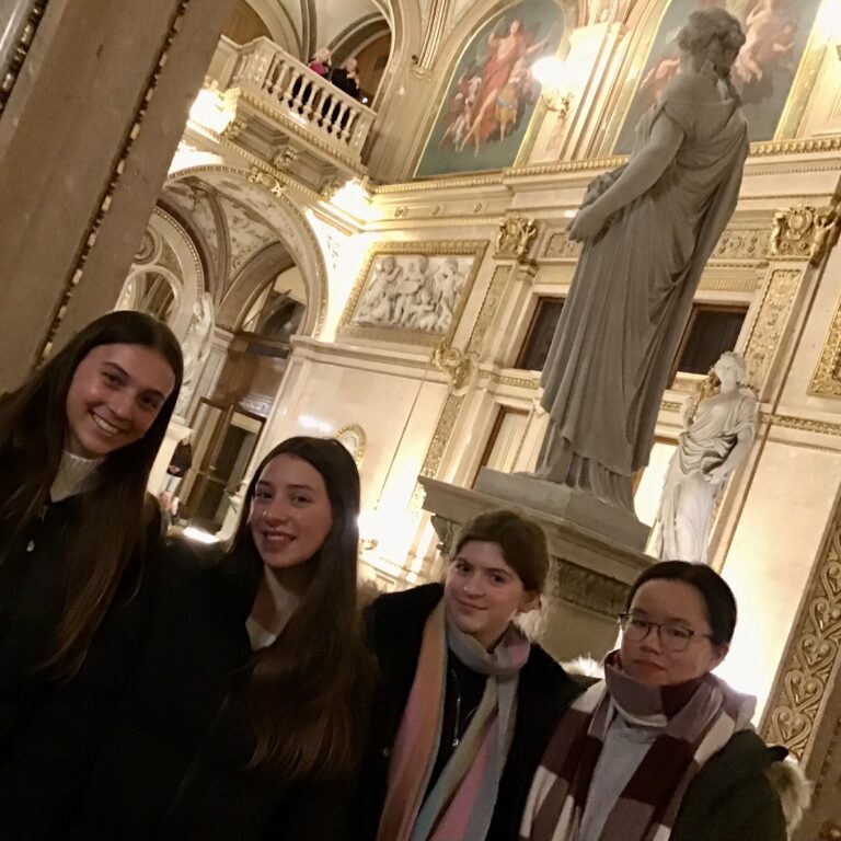Group of smiling young adults posing for a photo in a gilded, ornate museum hall with statues and murals.