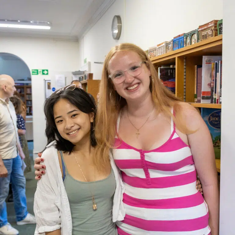 Two young women smiling for the camera in a library or bookstore, with shelves and people in the background.
