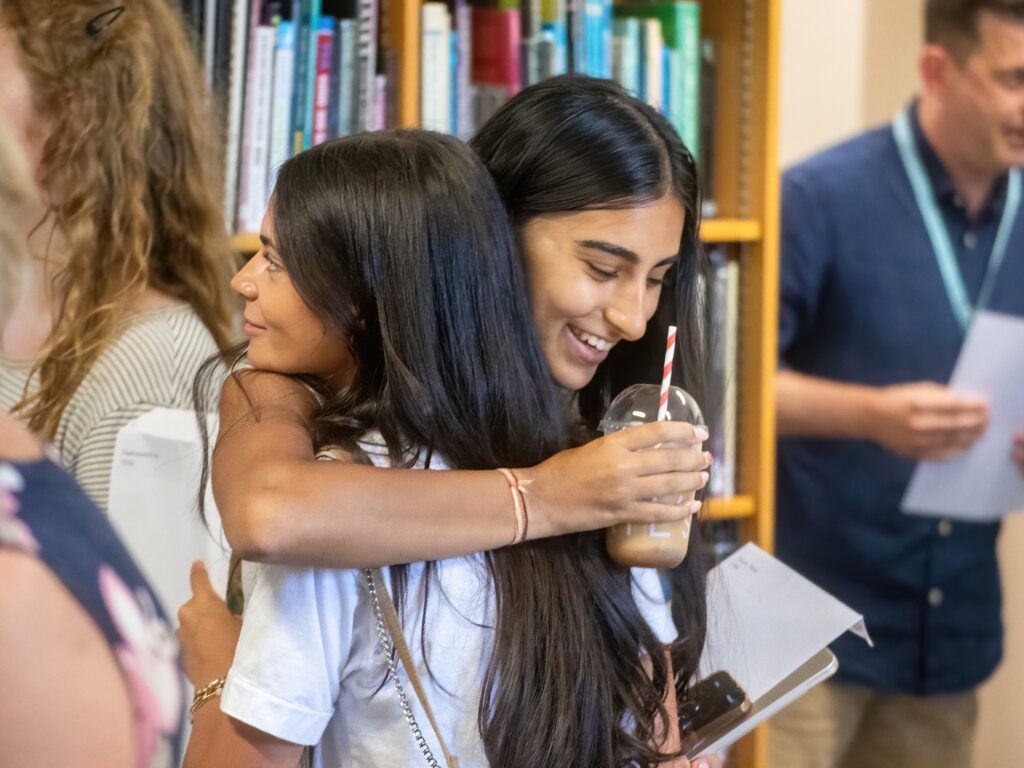 Smiling woman with long black hair hugging a friend in a library, holding a drink with a striped straw.