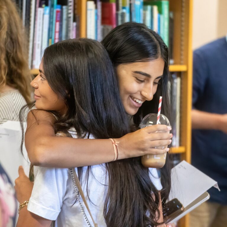 Smiling woman with long black hair hugging a friend in a library, holding a drink with a striped straw.