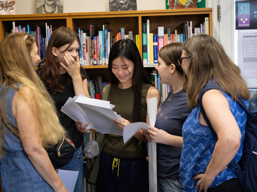 Group of five young women standing in a library, examining papers and talking together.