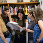 Group of five young women standing in a library, examining papers and talking together.