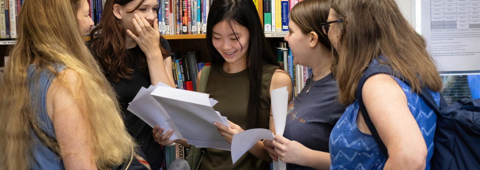 Group of five young women standing in a library, examining papers and talking together.