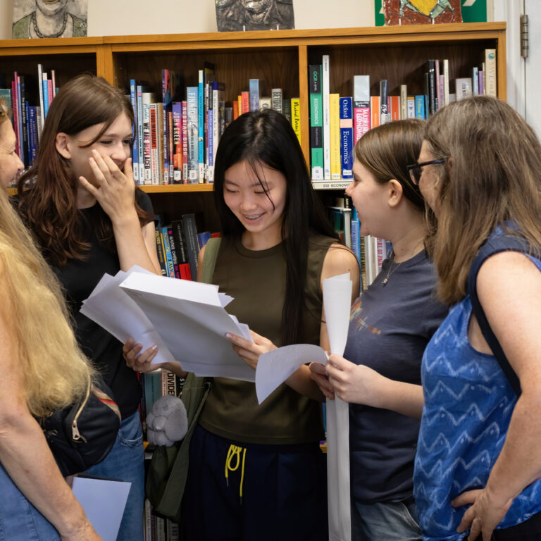 Group of five young women standing in a library, examining papers and talking together.